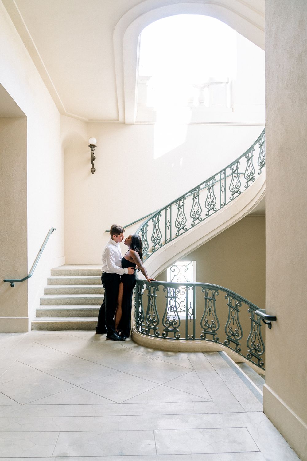 A couple embrace on an elegant marble staircase with ornate turquoise wrought iron railings in a bright interior.
