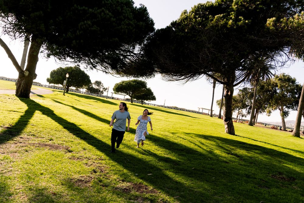 Two people in light clothing walk together on a grassy hillside overlooking the water on a sunny afternoon.