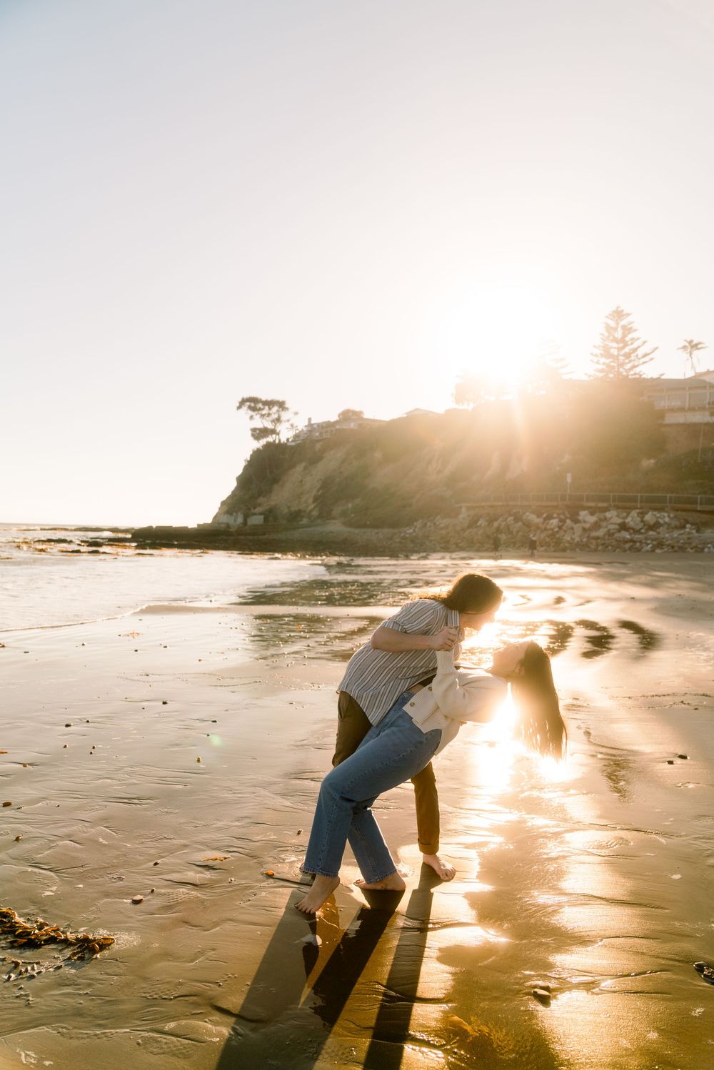 Two figures embrace on a sunlit beach at sunset with cliffs and ocean waves in the golden light.