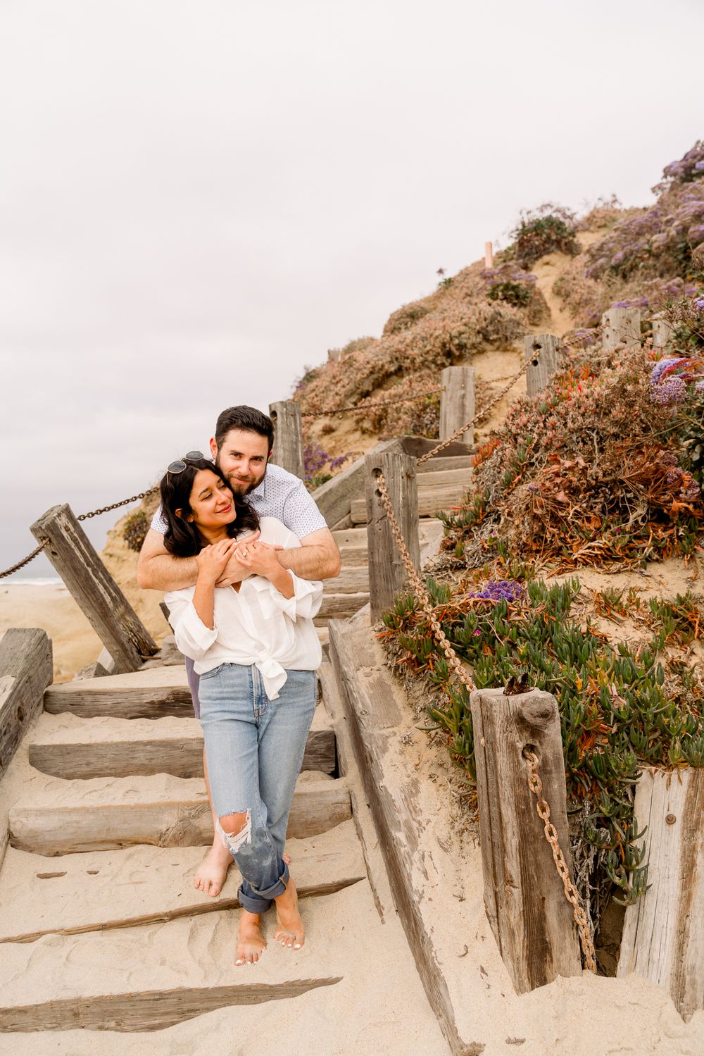 A couple embraces on rustic wooden beach stairs surrounded by coastal plants and flowers on a cloudy day.