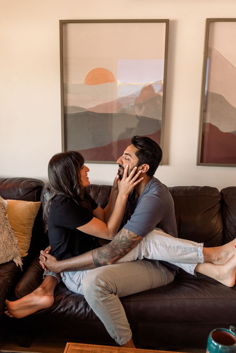 A couple shares an intimate moment on a brown leather couch with minimalist artwork on the wall behind them.