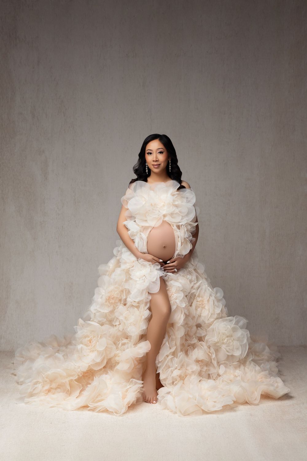 Pregnancy photoshoot featuring elegant cream gown with flowing ruffles and delicate lace details against neutral backdrop.