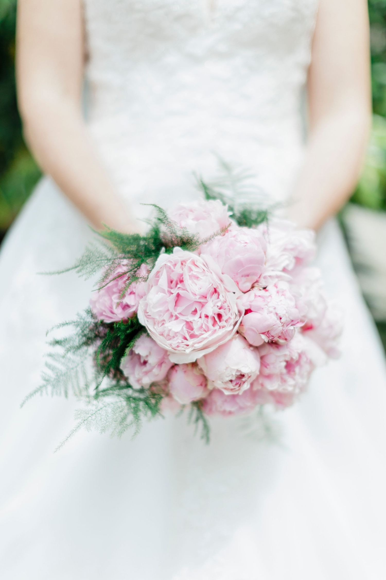 A delicate pink peony bridal bouquet with ferns against a white wedding dress.