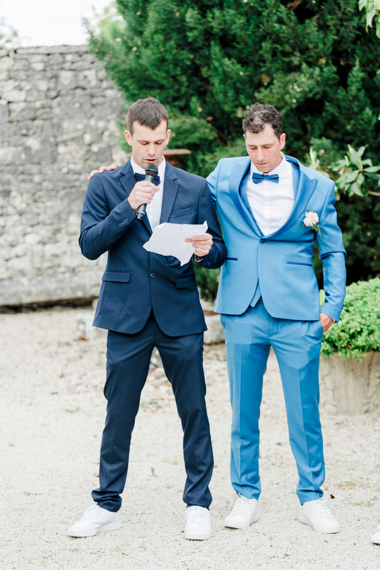 Two men in suits reading from papers during outdoor wedding ceremony.