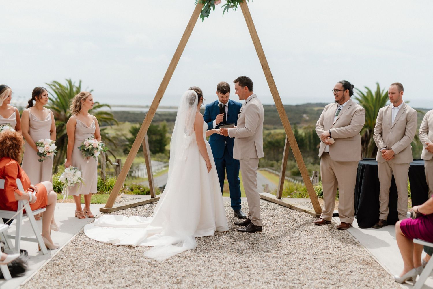 Outdoor beach wedding ceremony takes place under wooden arch with gravel ground and ocean view in background.