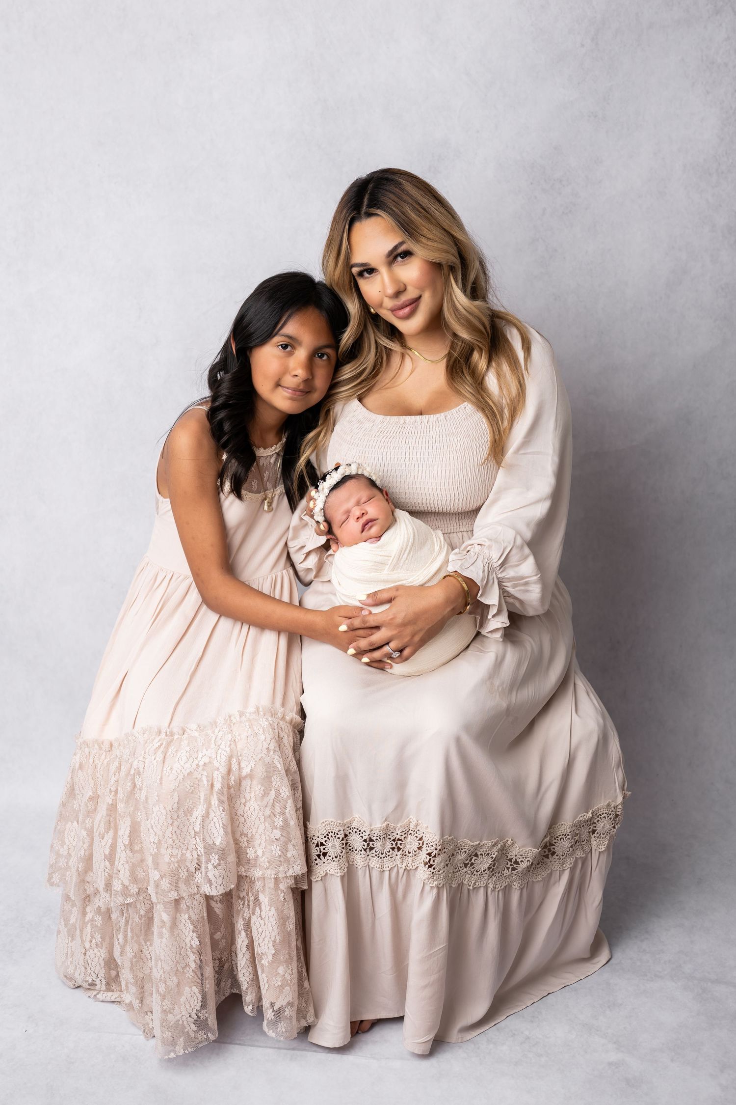 A tender portrait of two individuals in light-colored dresses holding a sleeping newborn baby against a grey backdrop.