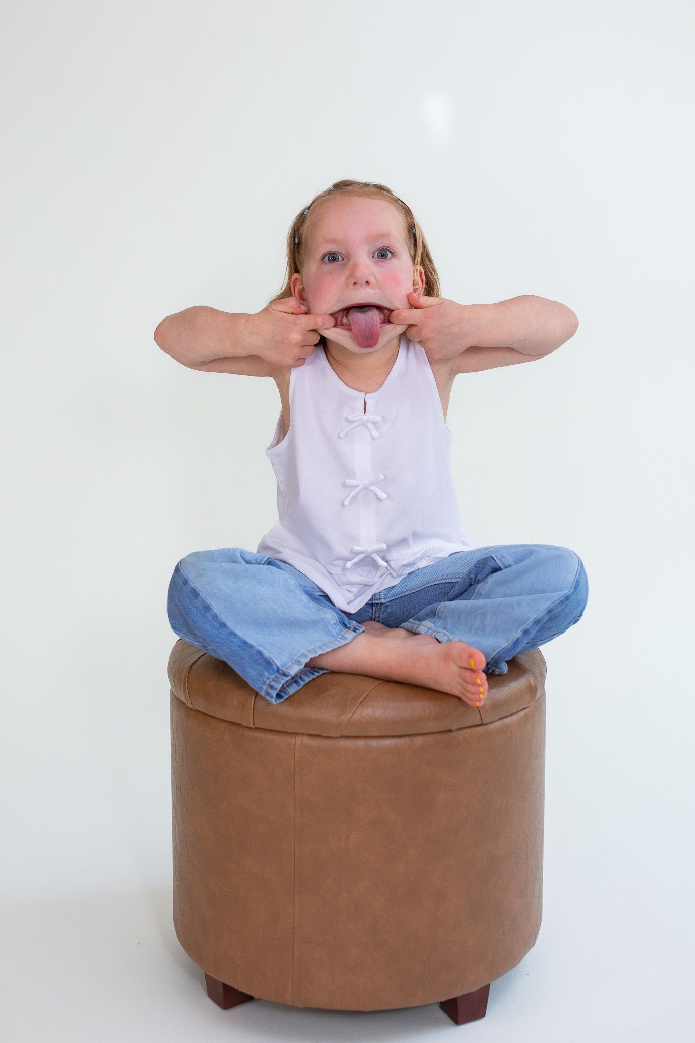 A child sits cross-legged on a brown leather ottoman while making a silly face and pulling their cheeks.