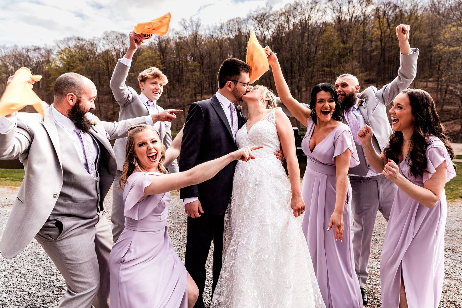 A wedding party celebrates outdoors wearing purple bridesmaid dresses and gray suits, waving orange napkins in the air.
