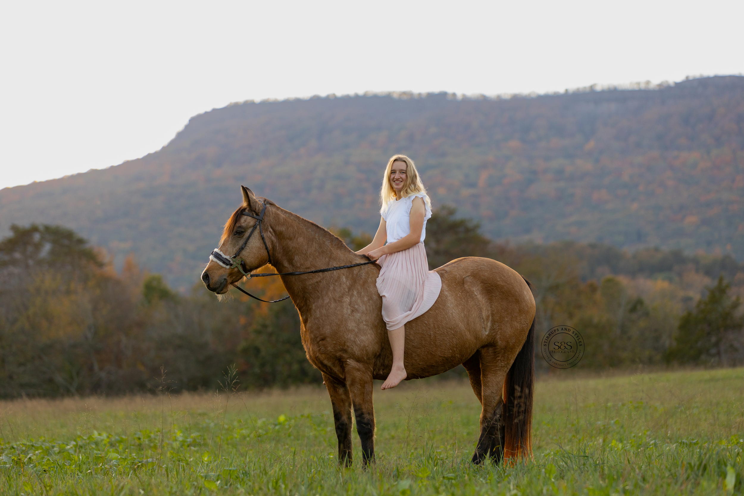 Rider in white dress sits atop brown horse in mountain meadow at sunset.