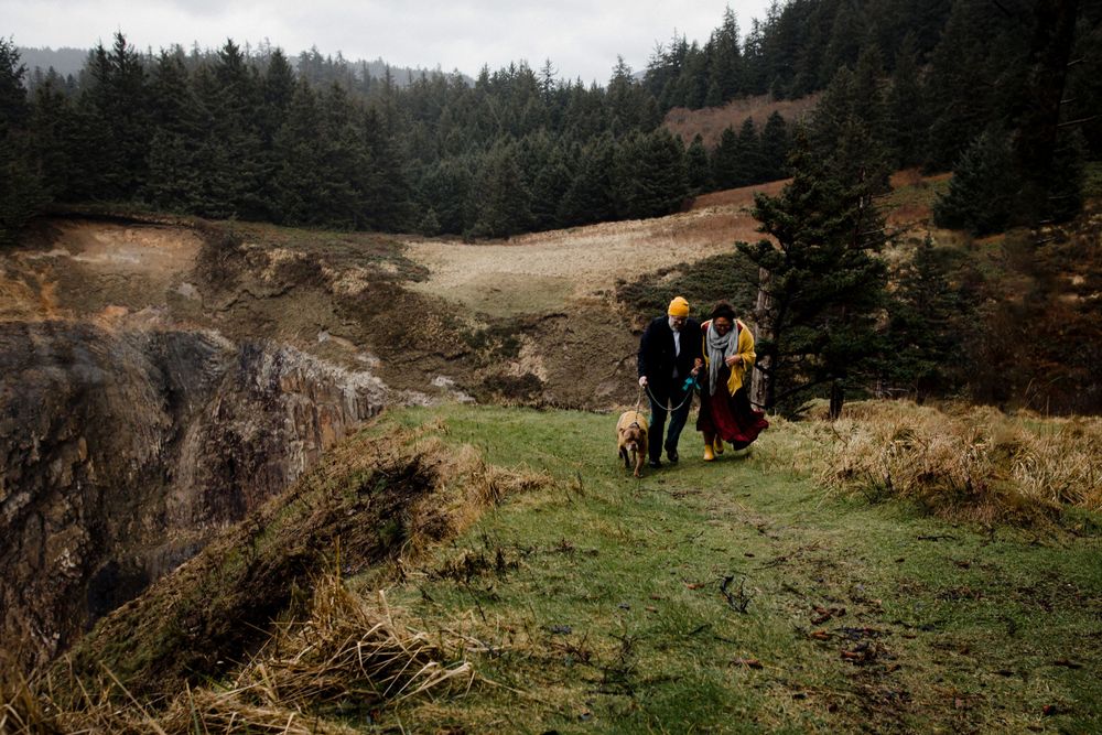 Two hikers with backpacks explore a scenic cliff edge surrounded by evergreen forest on a cloudy day.