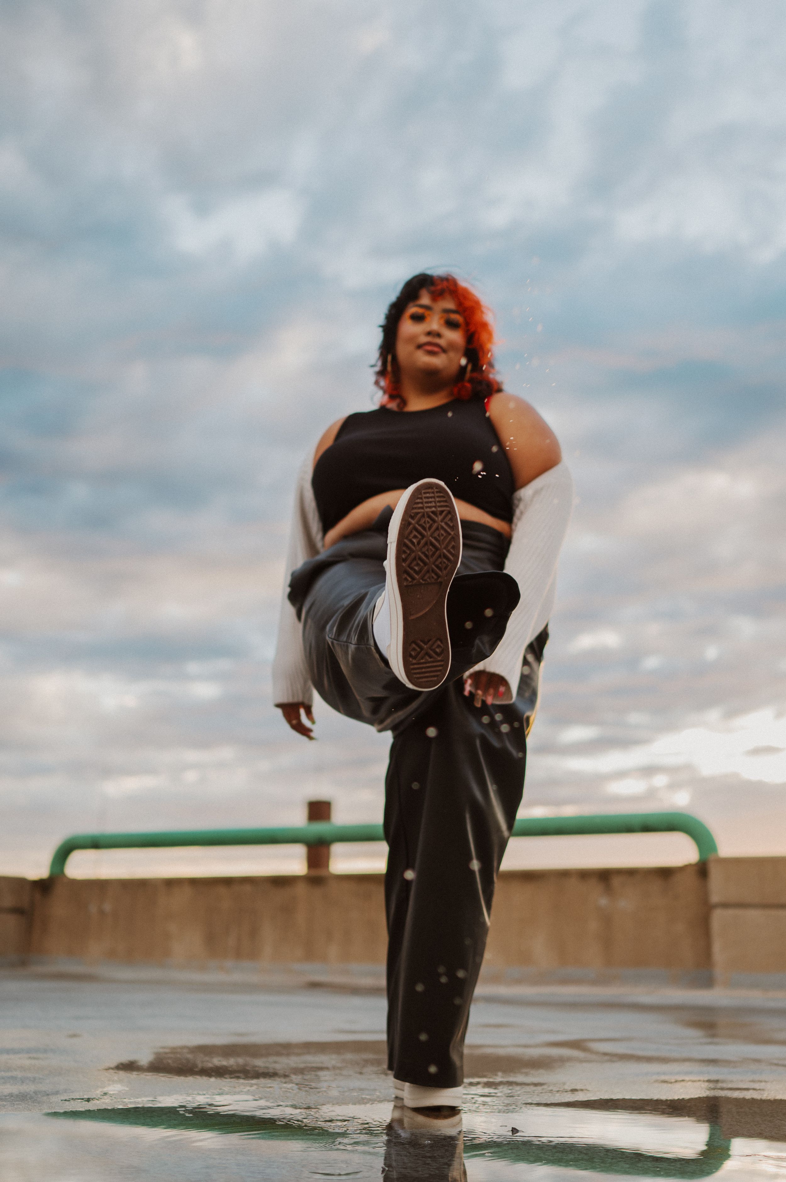 A dance pose on a rooftop at sunset shows a high kick against a cloudy sky backdrop.