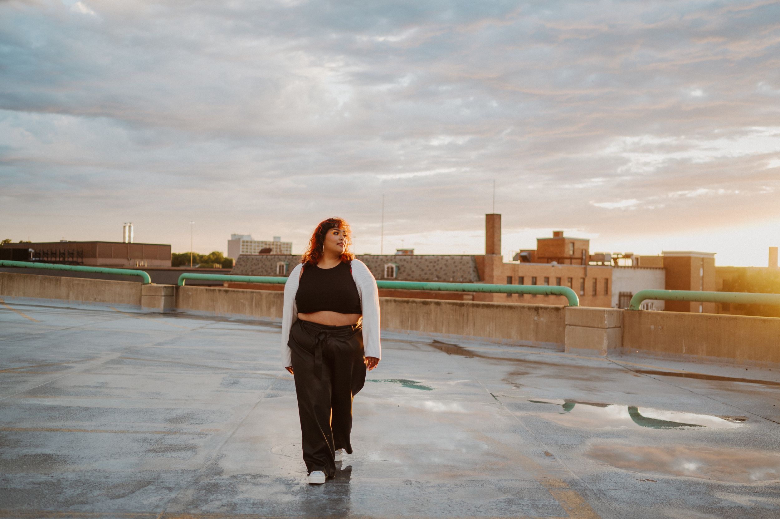 A person stands on a rooftop at sunset with a cityscape backdrop and reflective puddles of water on the surface.