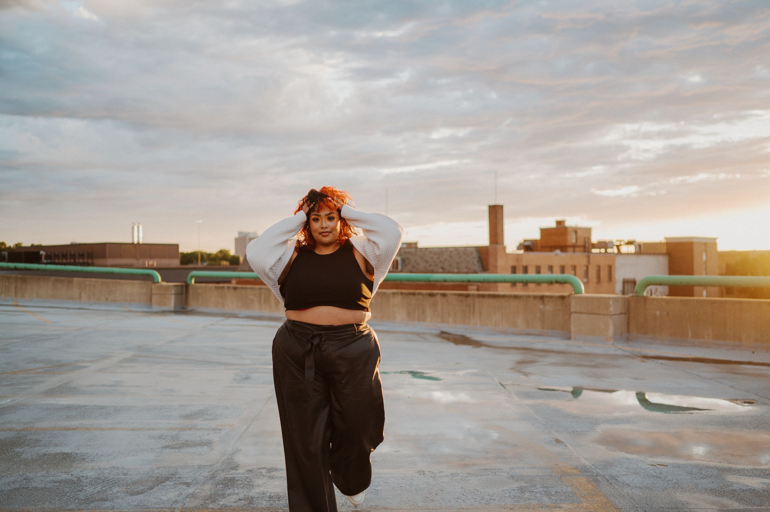 Someone in all black clothing poses on an urban rooftop at sunset with cityscape in the background.