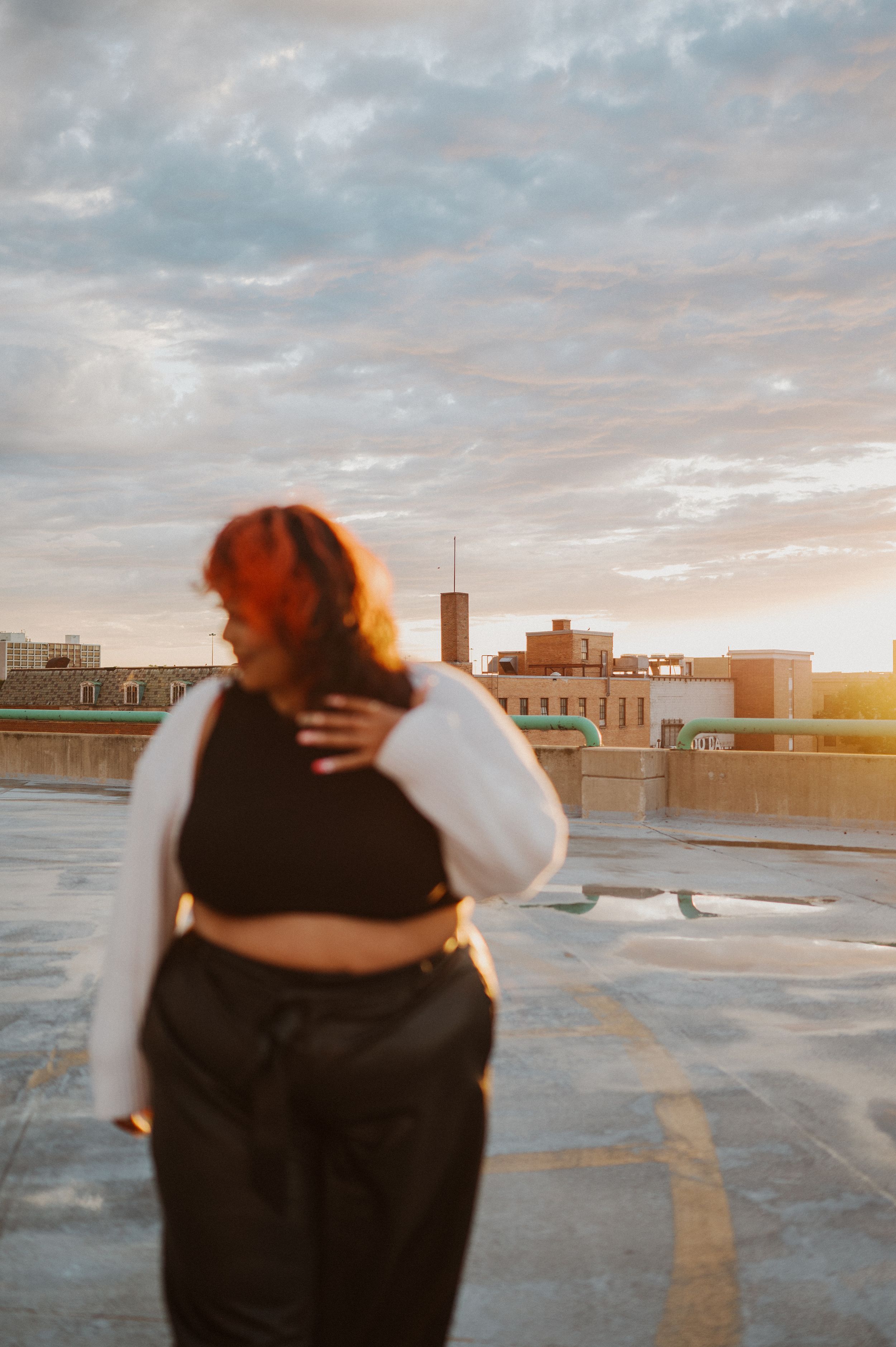 A plus-size figure stands on a rooftop at sunset wearing black clothing against an urban cityscape backdrop.