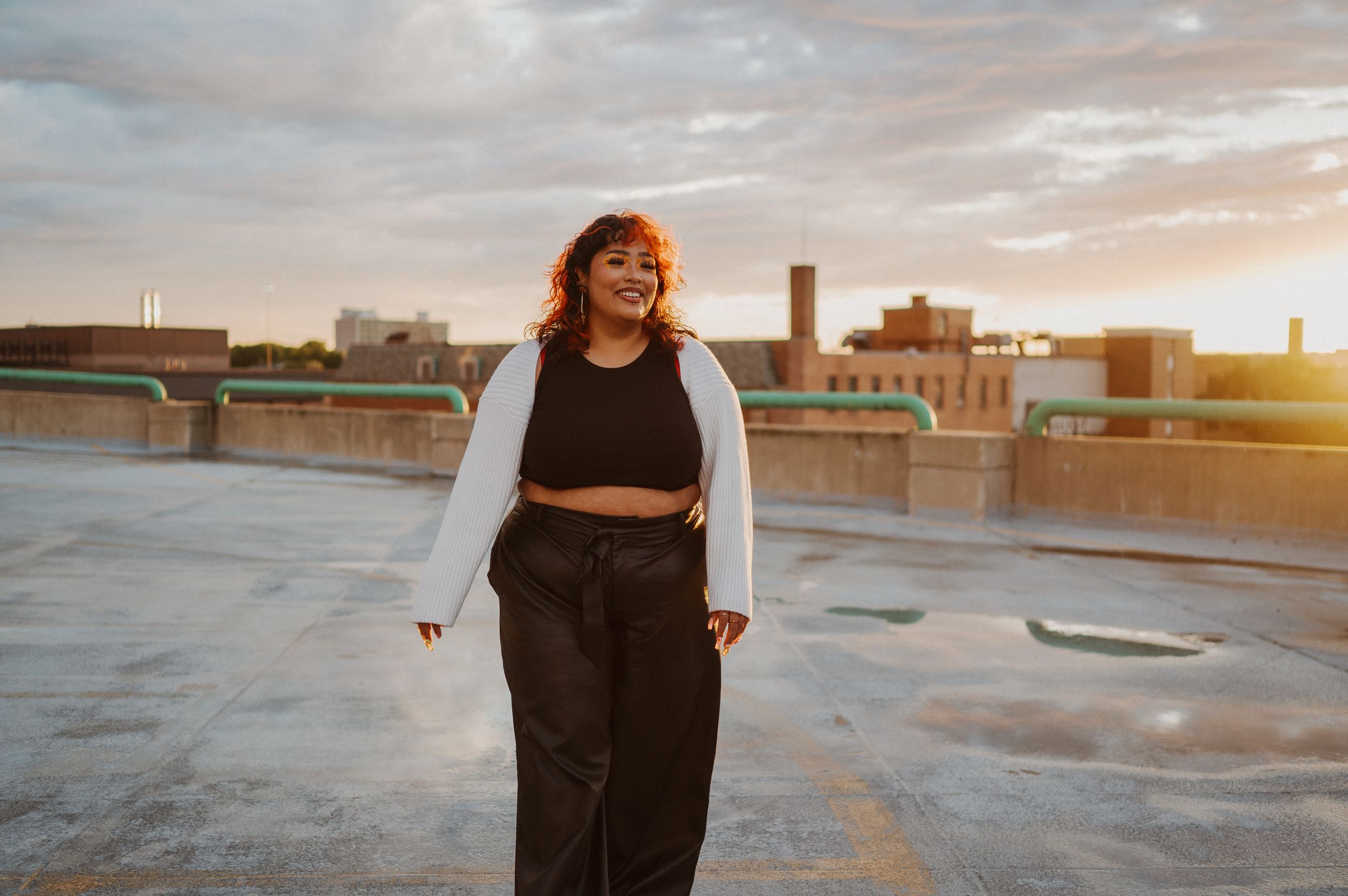 A person in a black outfit and white cardigan stands on a rooftop at golden hour with urban buildings in the background.
