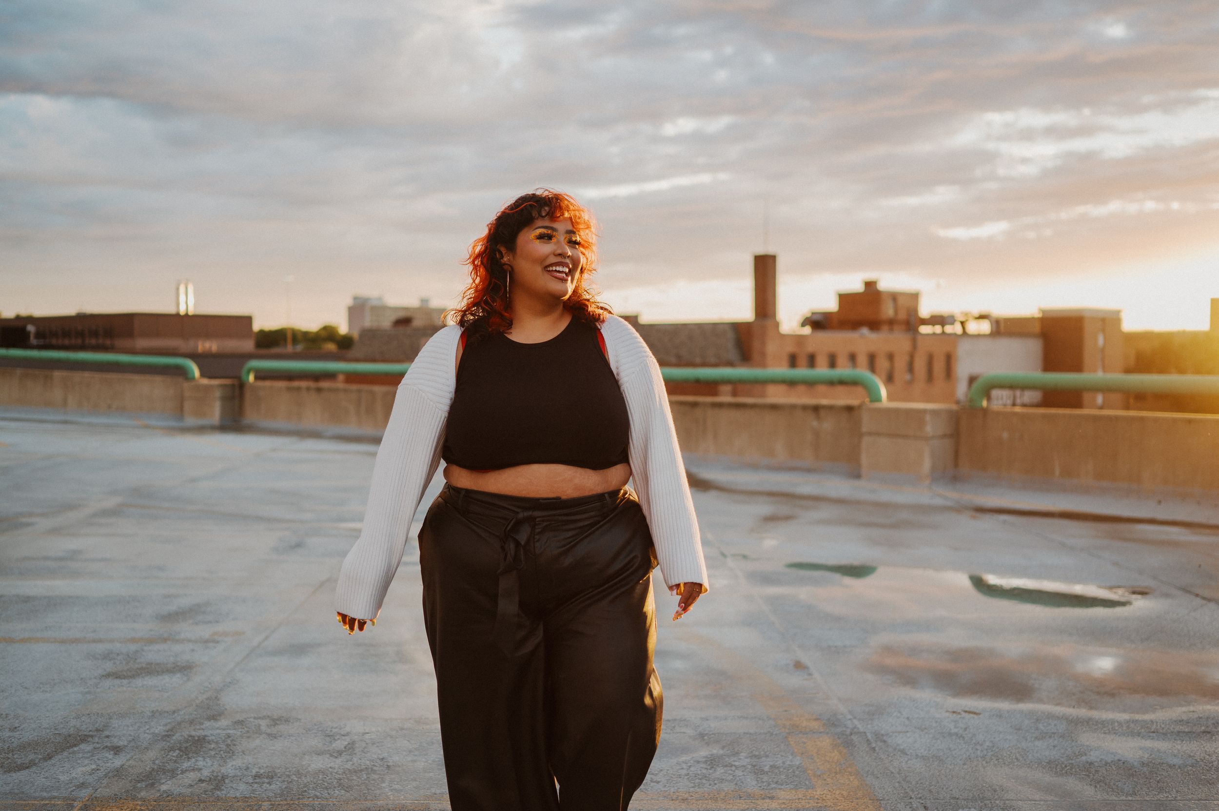 A person wearing a white cardigan and black crop top poses on an urban rooftop during golden hour sunset.