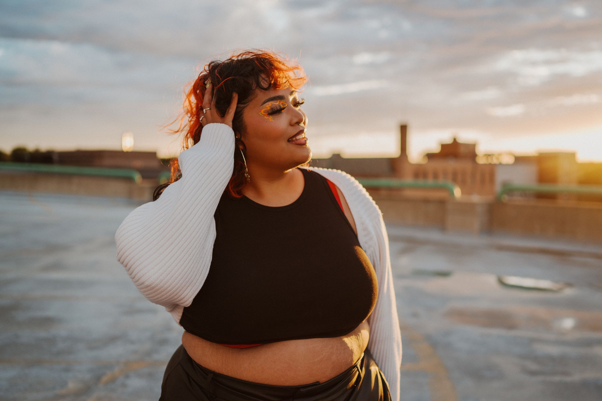Person wearing a black top and white cardigan poses on rooftop during golden sunset with cityscape in background.