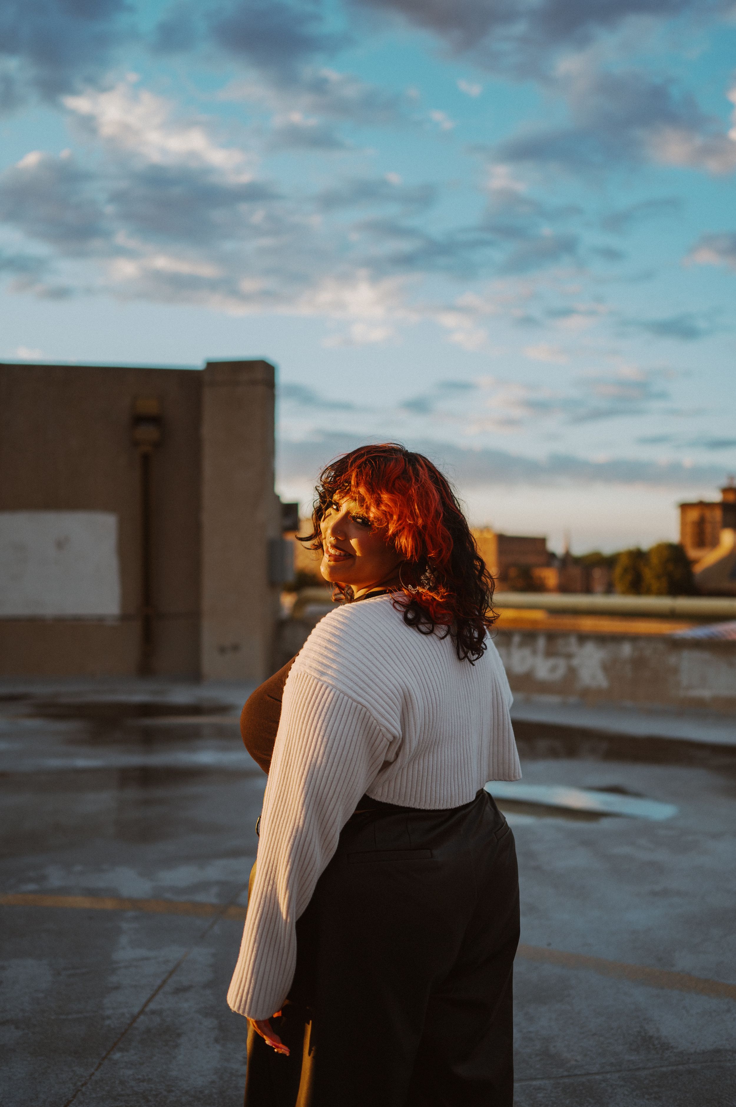 A figure in a white sweater gazes at the sunset over a rooftop under dramatic cloudy skies.