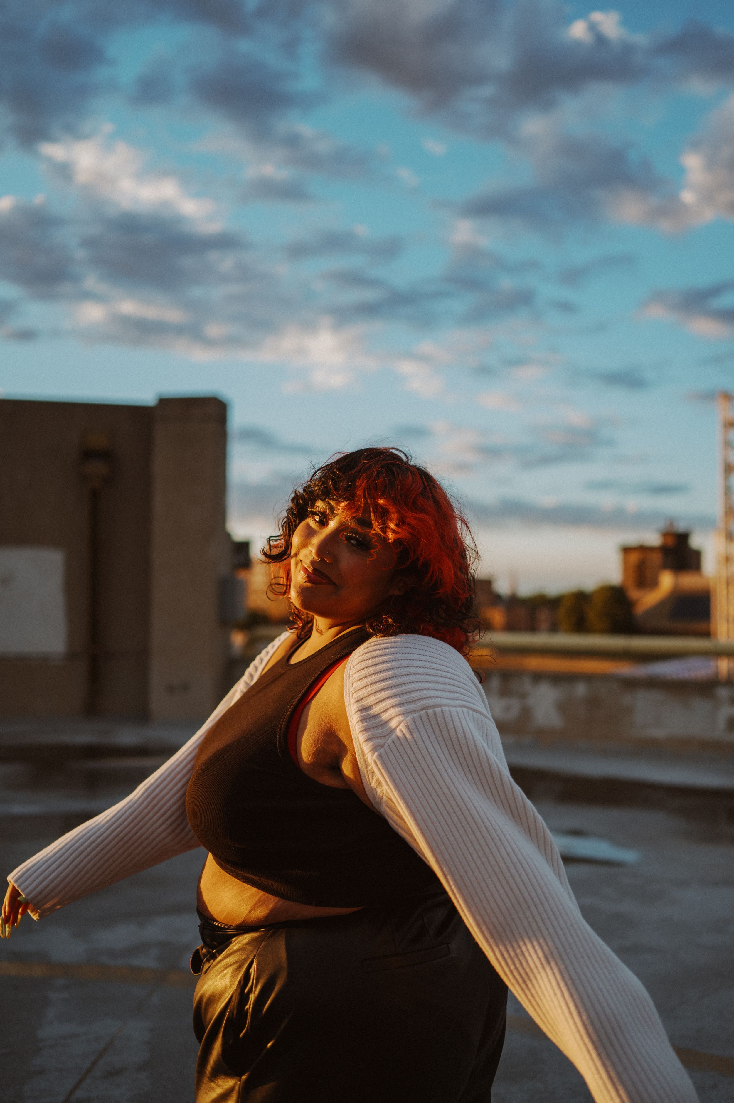 A joyful dance pose captured during sunset on a city rooftop with dramatic clouds in the background.