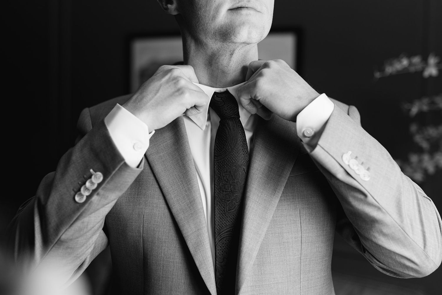 A close-up black and white photo of someone adjusting a tie with a suit jacket.