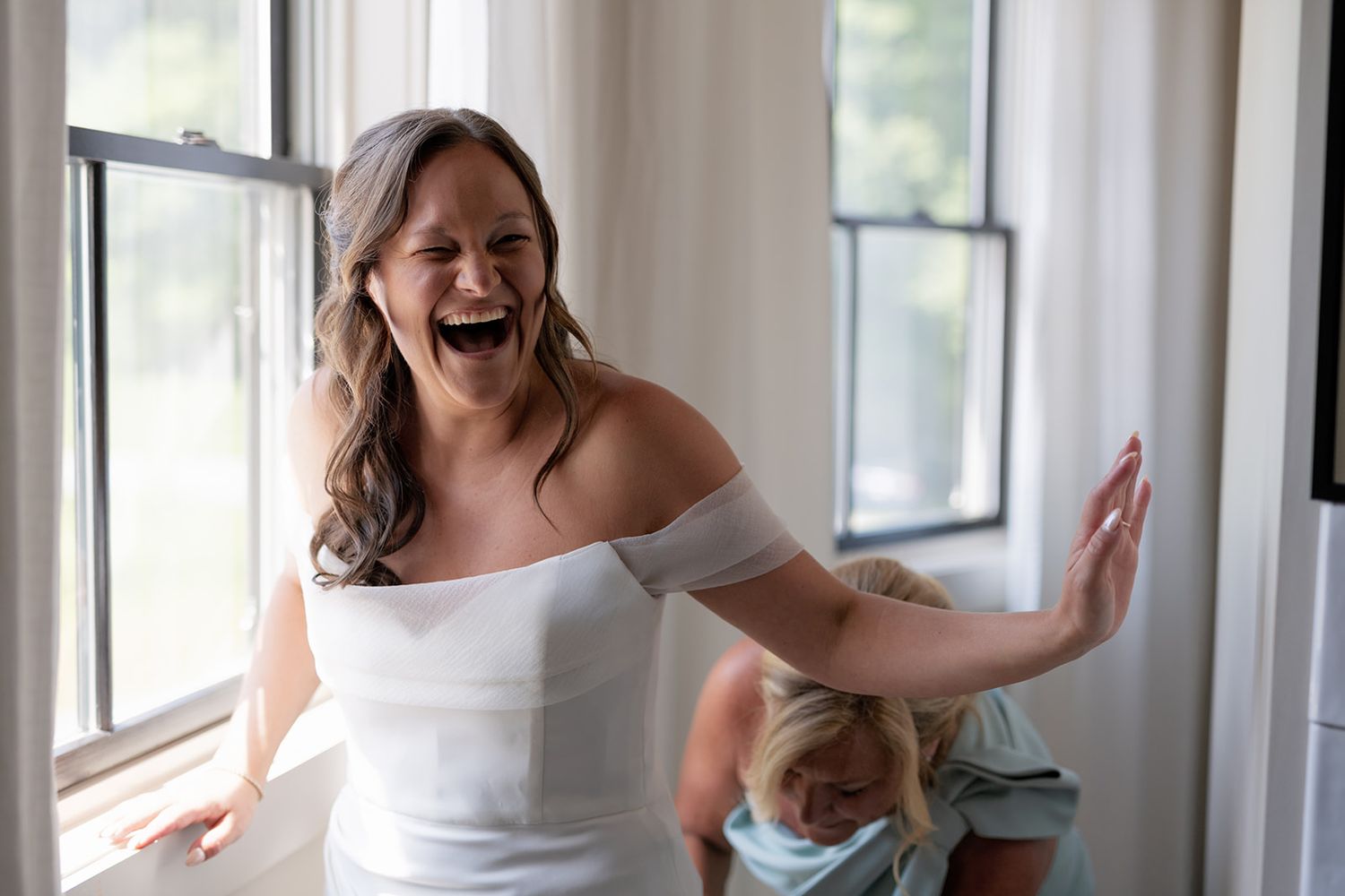 A joyful bride in a white off-shoulder wedding dress laughs heartily near sunlit windows.