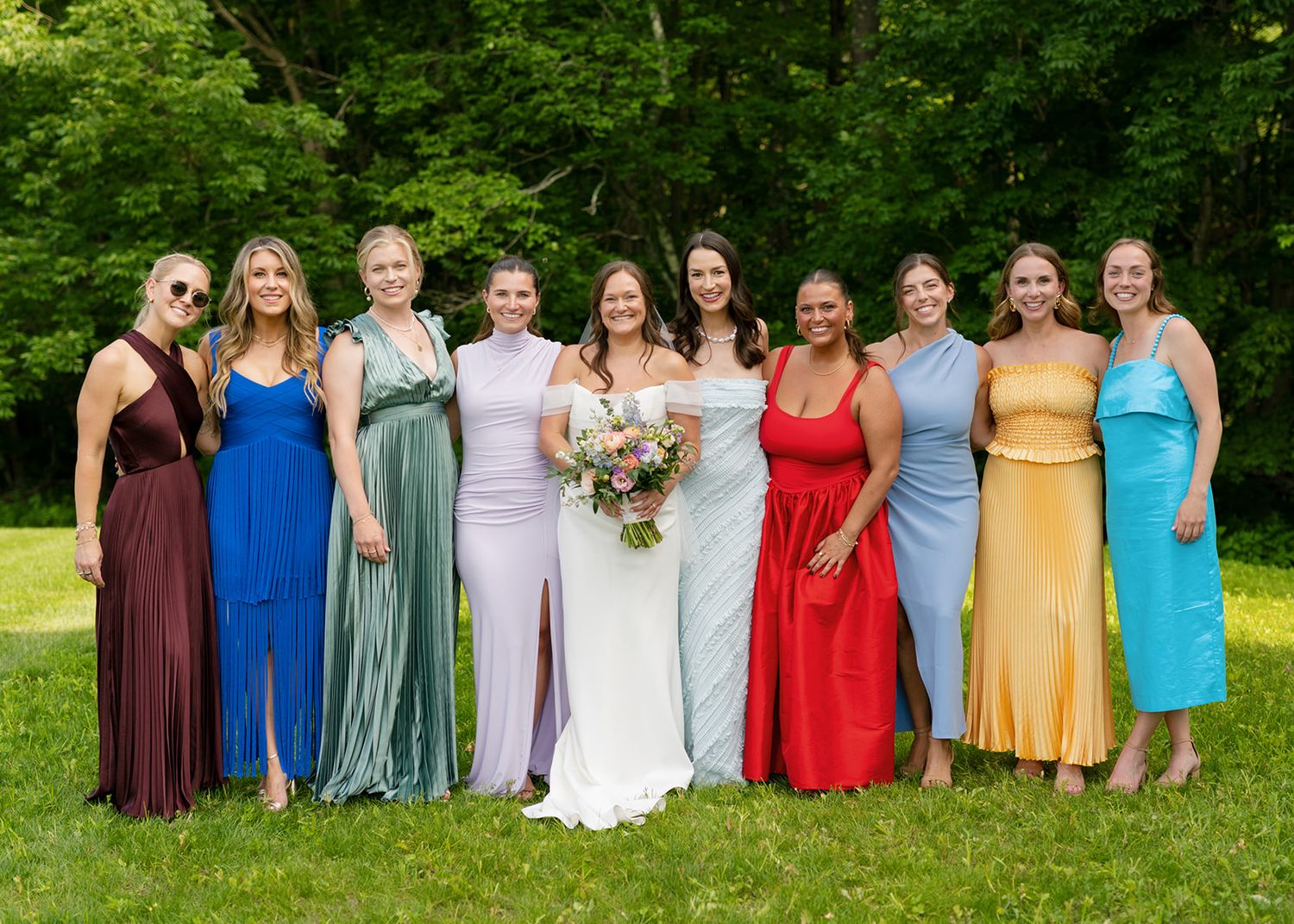 Wedding party in colorful bridesmaid dresses of burgundy, blue, sage, pink, red, yellow and aqua pose outdoors on green lawn.