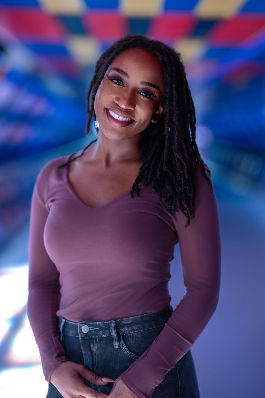A woman poses in a burgundy long-sleeve top and jeans against a blue and pink illuminated backdrop for a photo series.