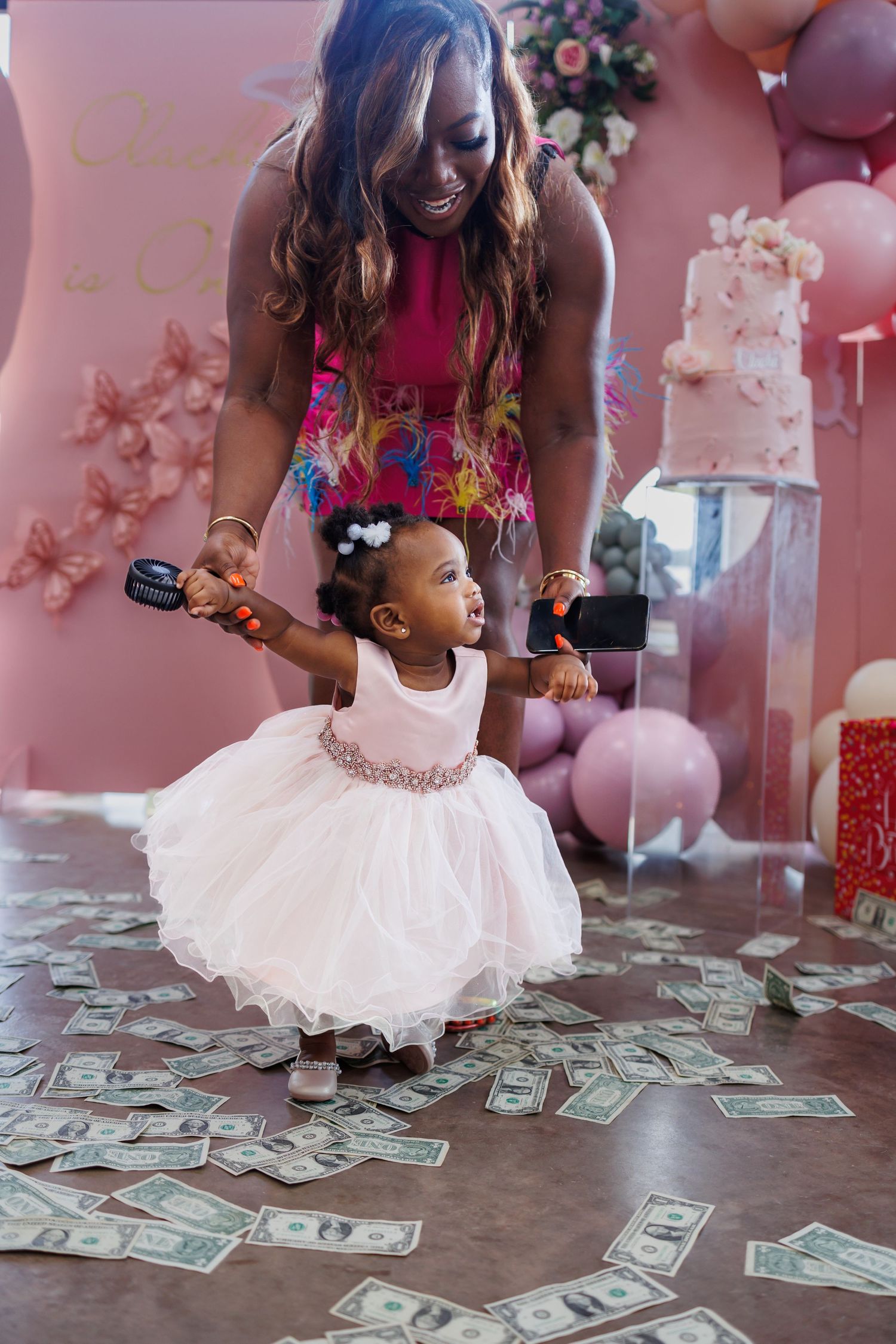 Mom in a bright pink dress helps a little girl in a tulle dress stand at her first birthday party at Bella Cavalli