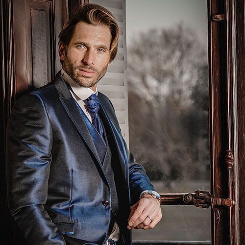 Man in navy blue suit stands elegantly by a window in a classic interior setting during a professional photoshoot.