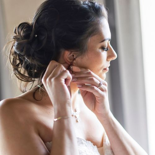 Elegant bride with dark updo hairstyle putting on earrings by window in white wedding dress.