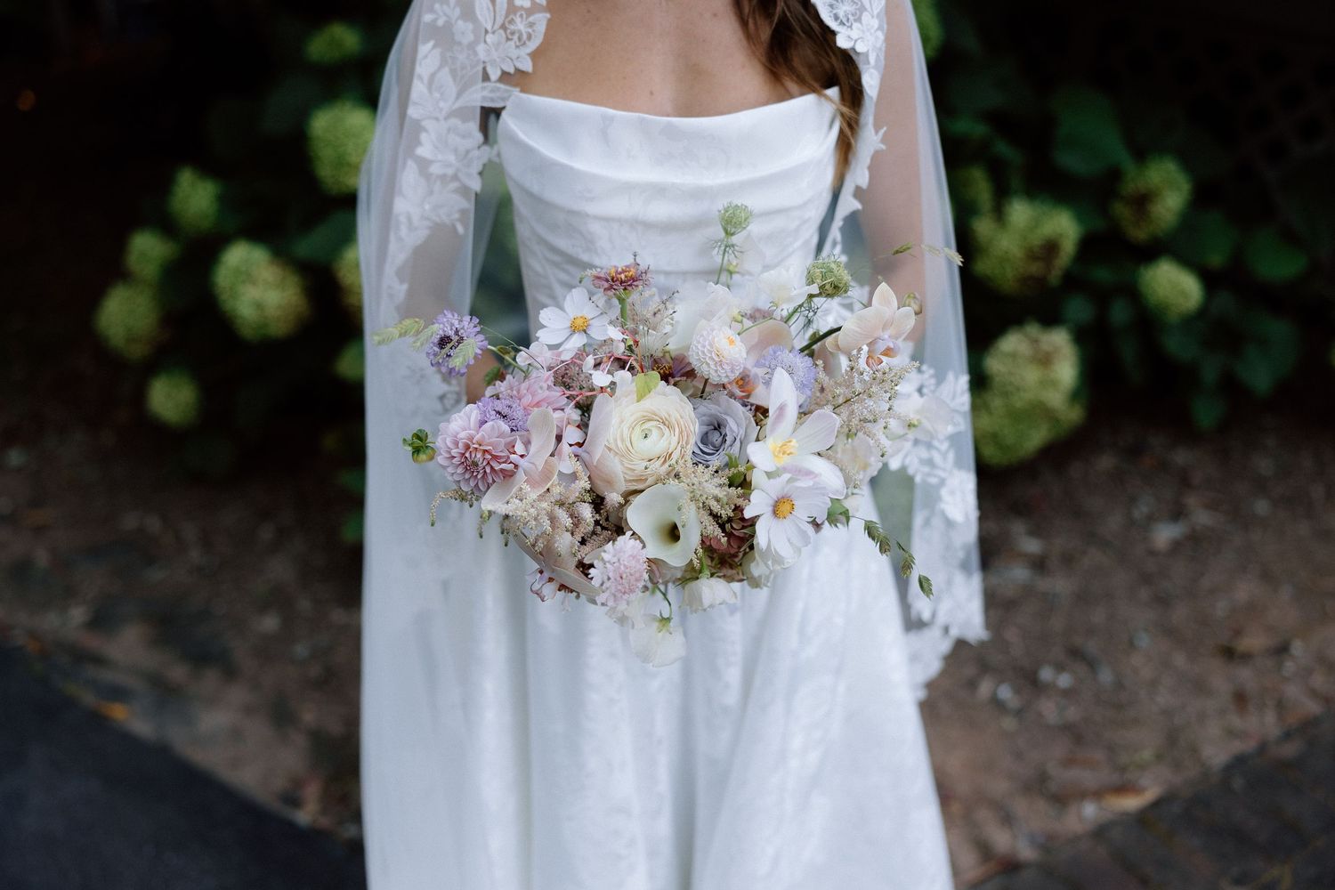 A delicate bridal bouquet of blush pink and white flowers is held against a white wedding dress with lace details.