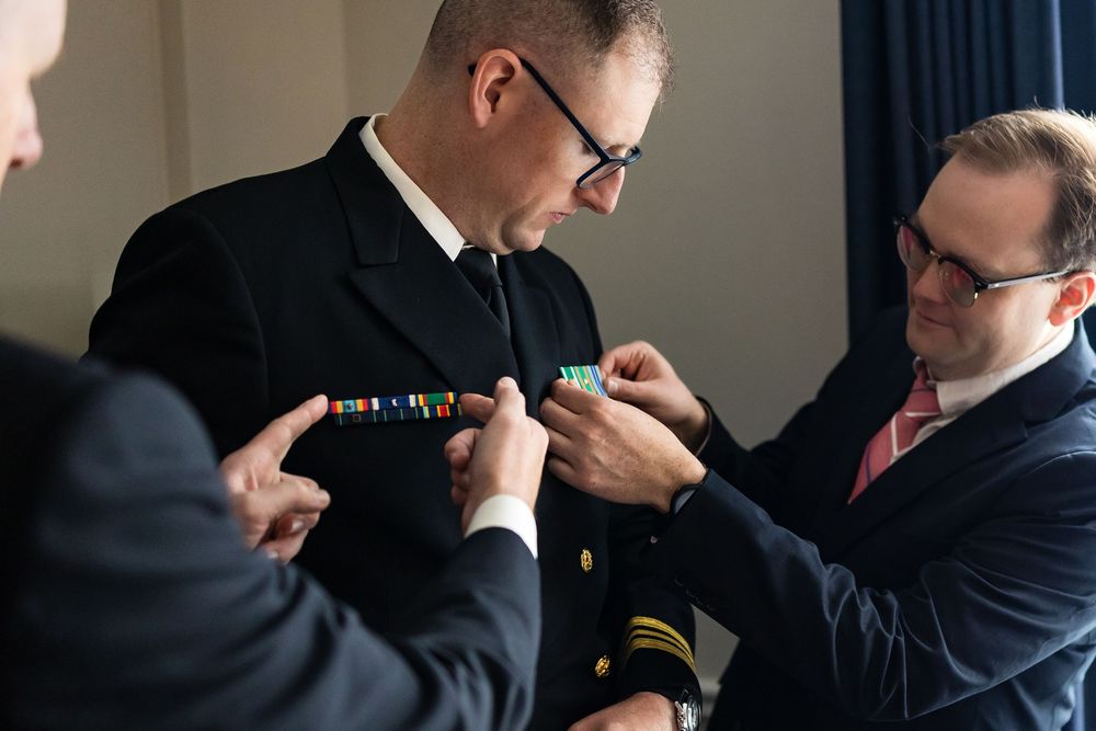 Two men in suits adjust a tie during formal wedding preparation.