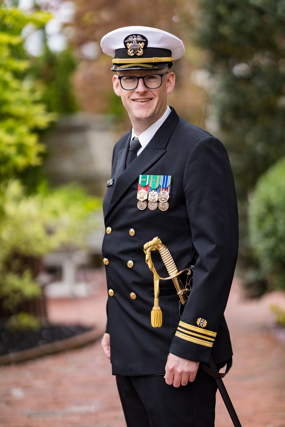 Naval officer in dress uniform with medals and sword standing outdoors.