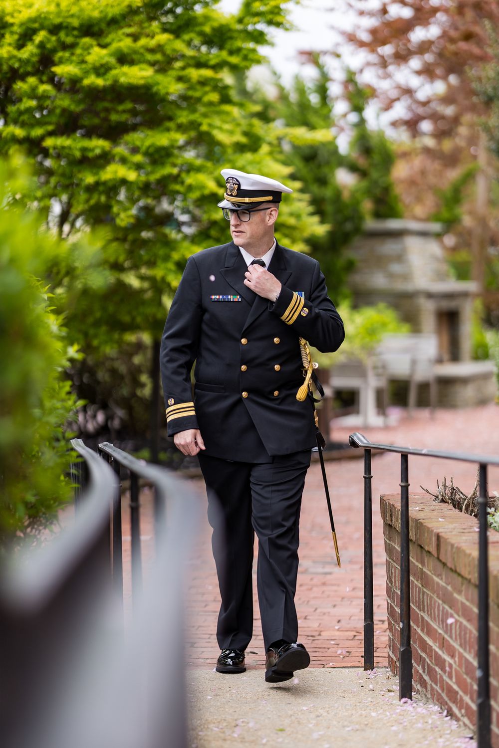 Naval officer in dress uniform walking along brick pathway in garden setting.