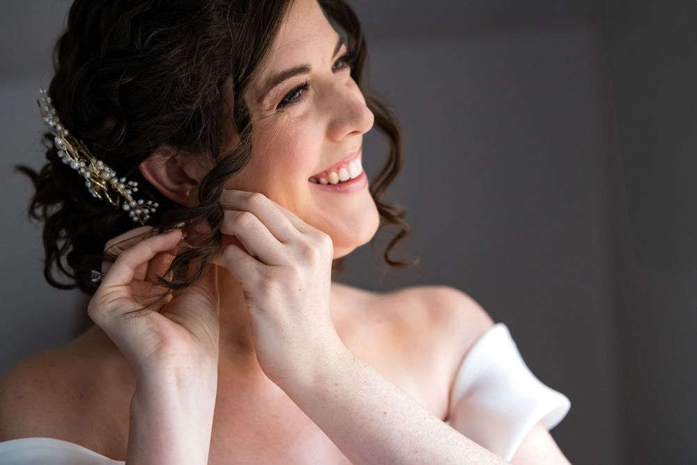 Joyful bride wearing white dress and pearl headpiece adjusting earrings.