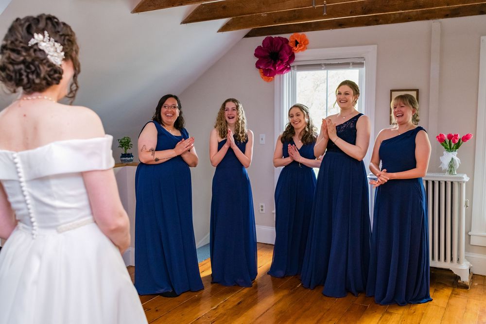 Bride in white dress reveals her gown to bridesmaids in navy blue dresses who react with joy.