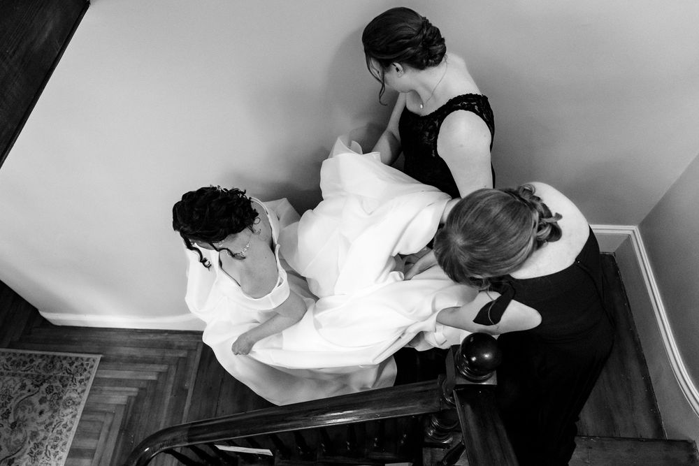 Black and white overhead view of bride in wedding dress ascending staircase with helpers.