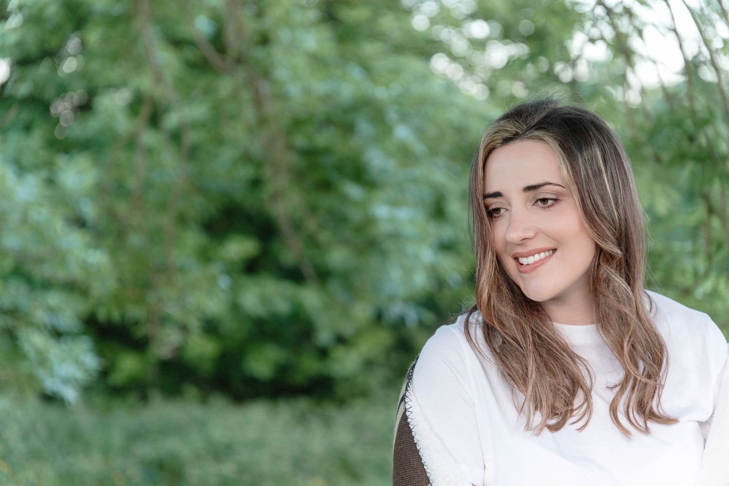 A person wearing a white sweater smiles outdoors with green foliage in the background.