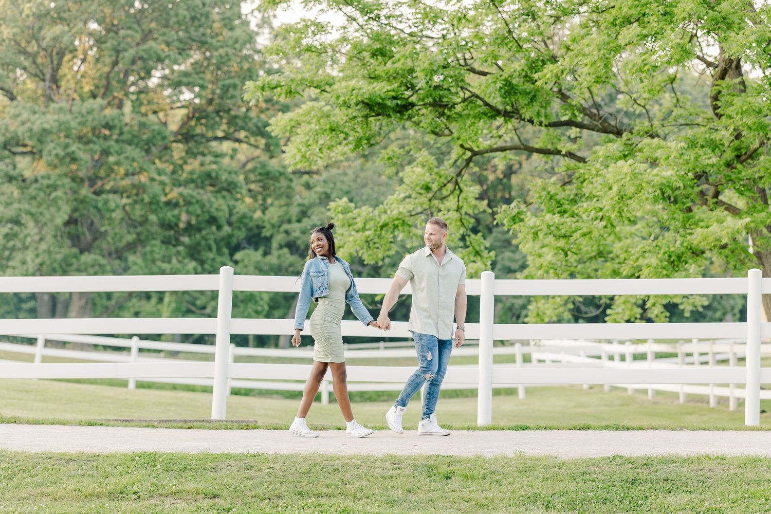 Two people enjoying a sunny day walk together along a white fence path surrounded by lush green trees and grass.