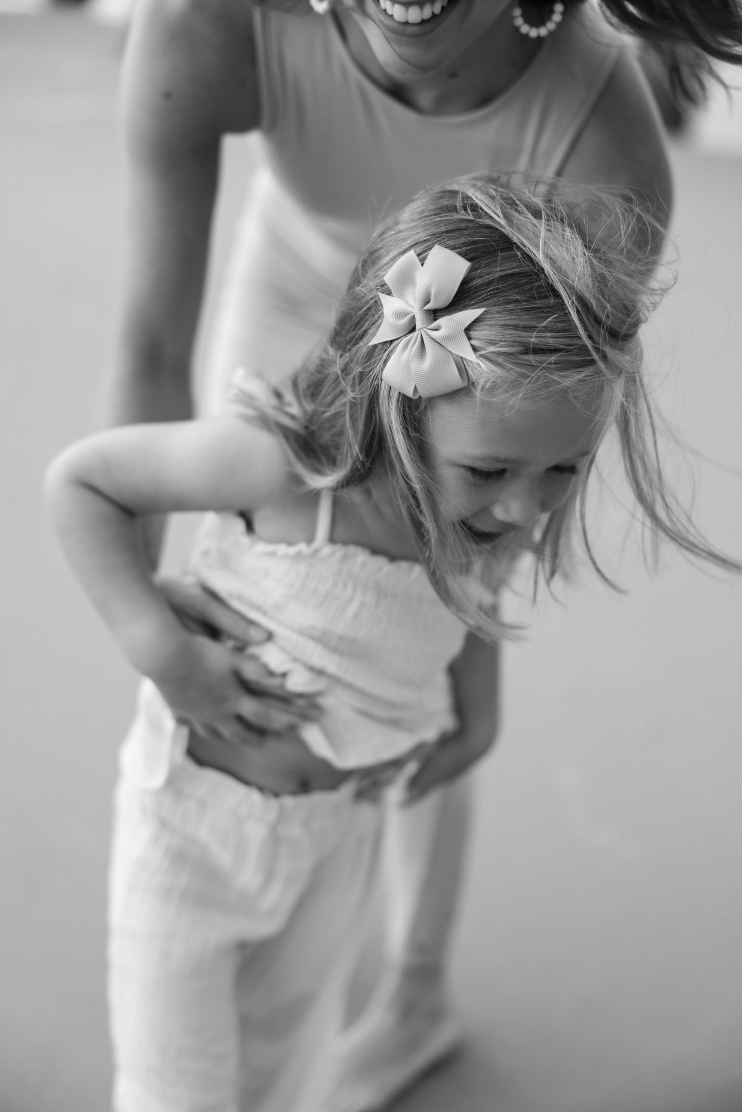 A black and white photo shows a joyful moment with a small child wearing a white dress and hair bow being tickled.