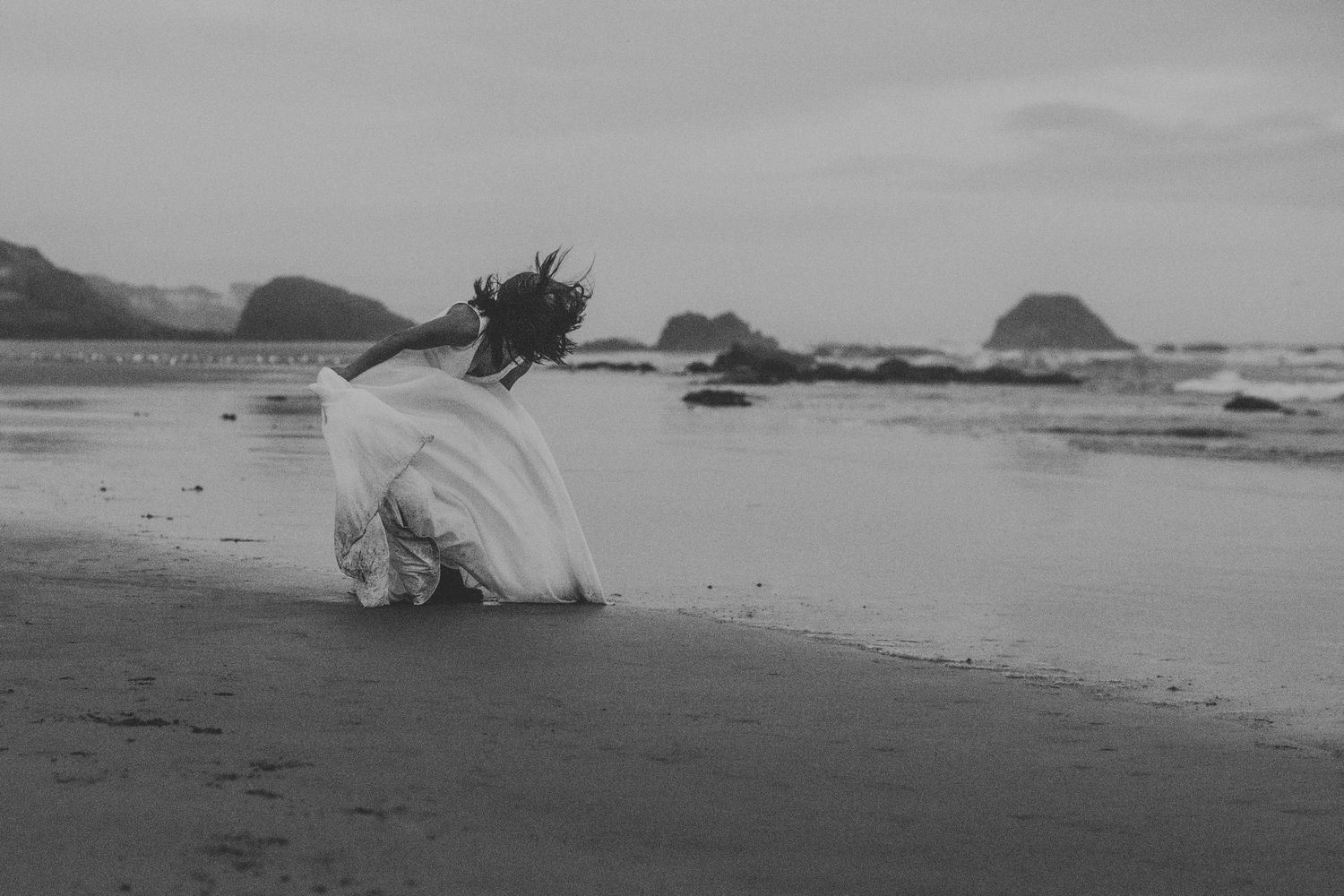 A person in white clothing sits alone on a misty beach with coastal rock formations in the distance.