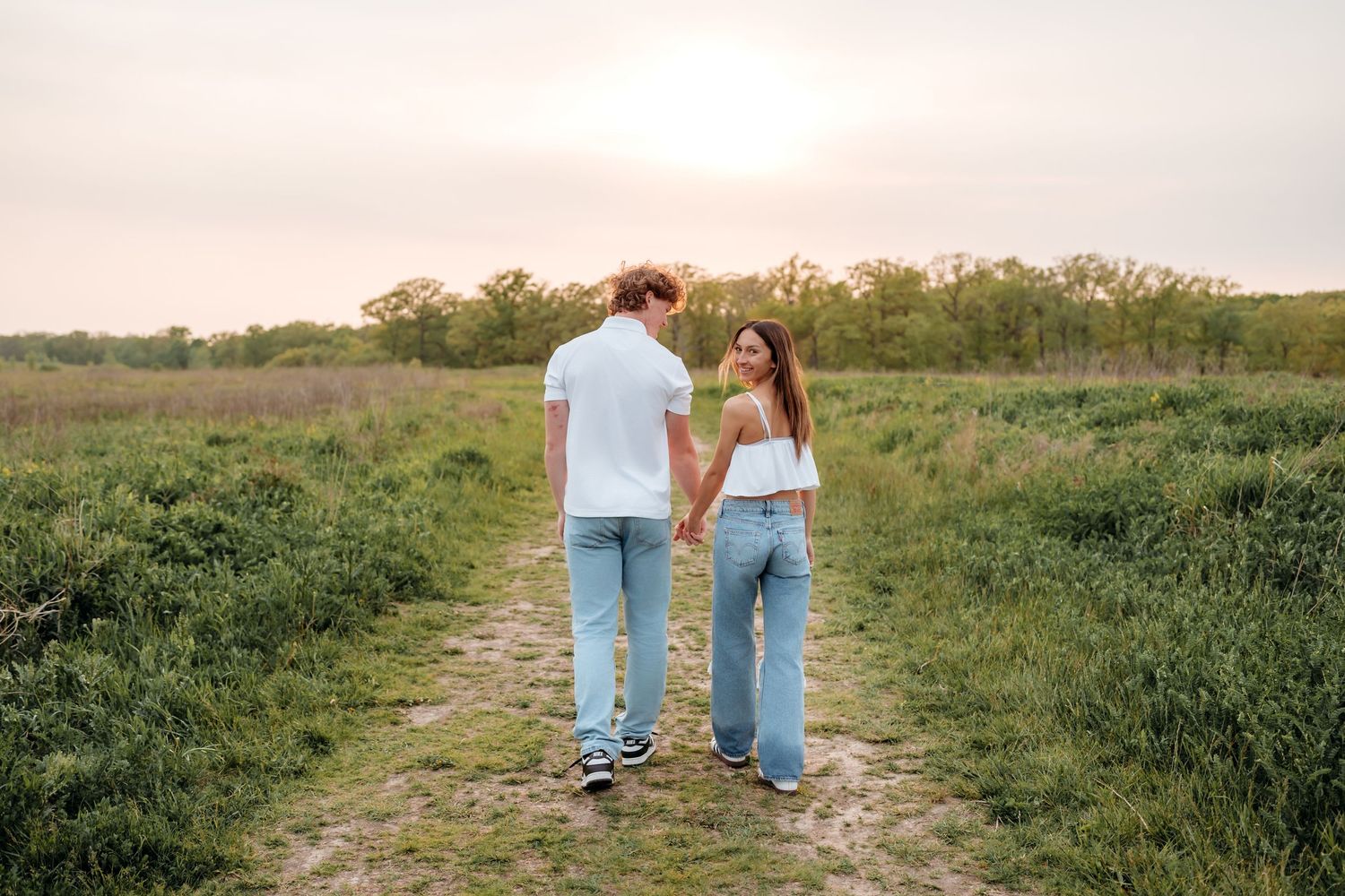 A couple in blue jeans walks hand in hand along a grassy path during golden hour.