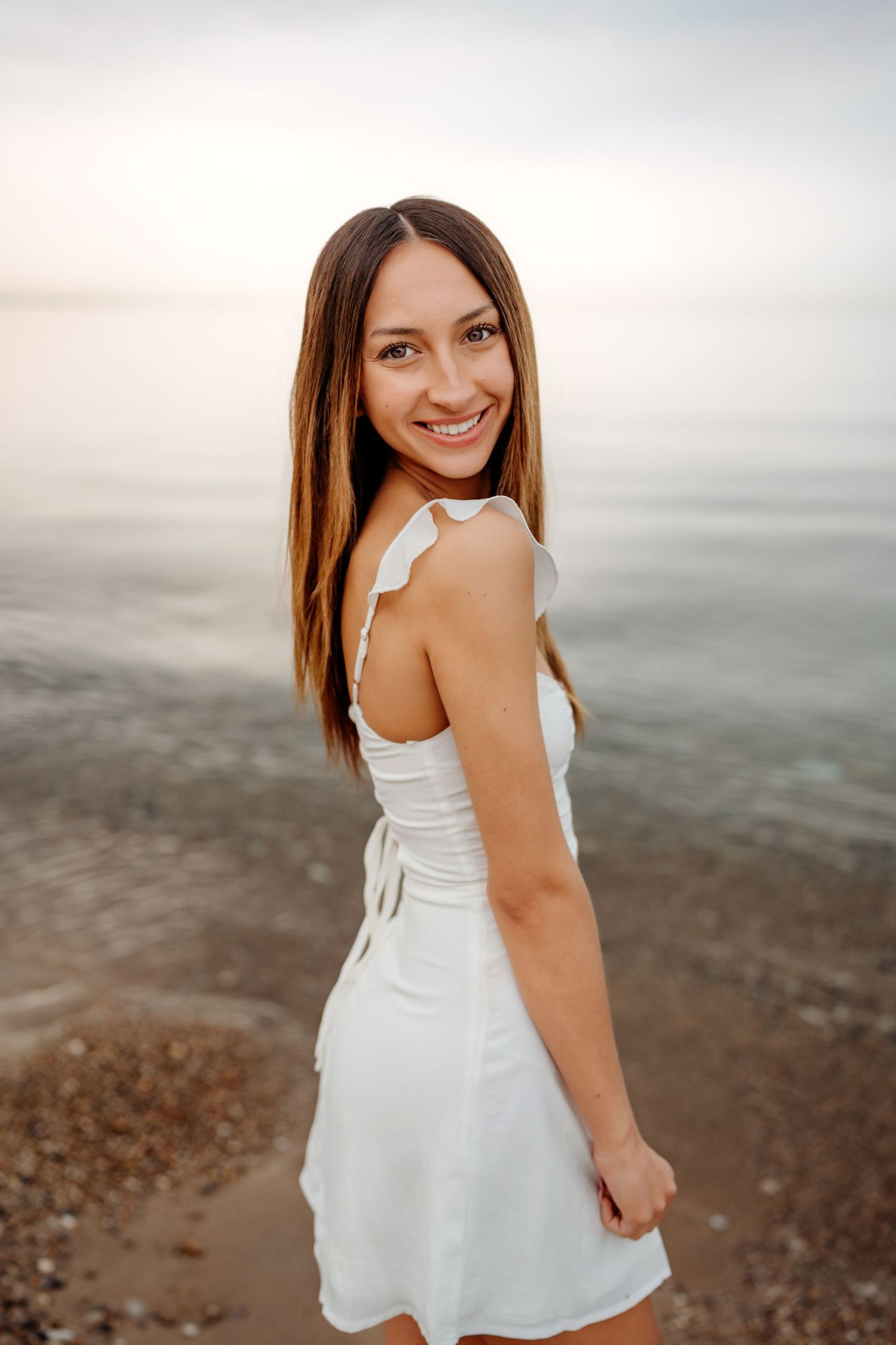 A person in a white summer dress poses by a misty beach at sunset.