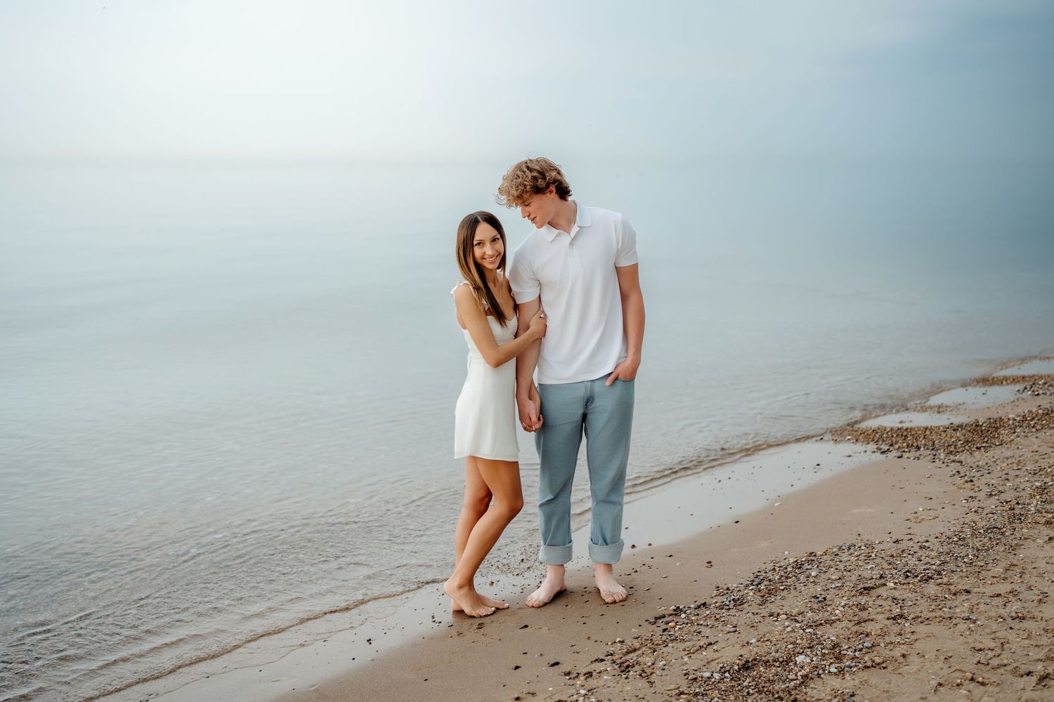 A couple in casual white outfits standing together on a peaceful beach during sunset.