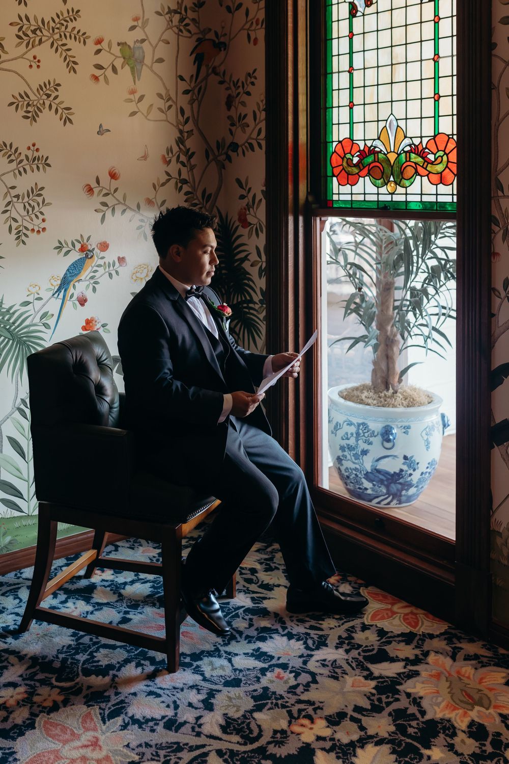 A well-dressed guest sits in an elegant room with floral wallpaper, stained glass window, and blue-and-white chinoiserie decor.