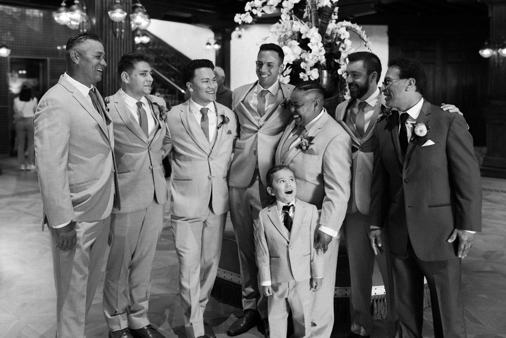 A wedding party in suits gathers together beneath decorations in an elegant black and white photograph.