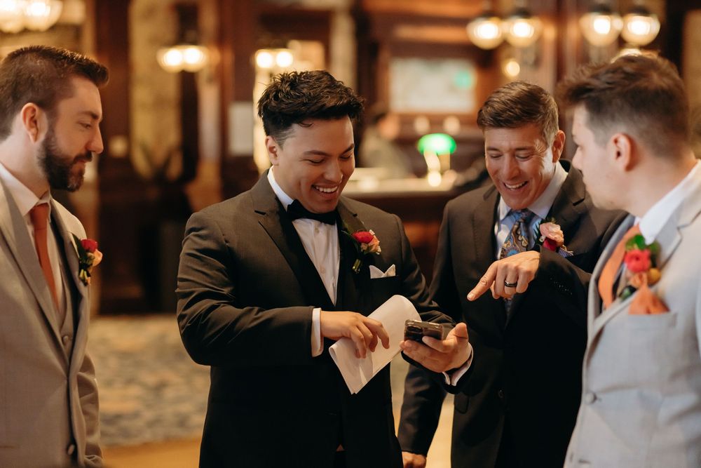 A group of groomsmen in suits and bowties gather to look at something on their phones before the ceremony.