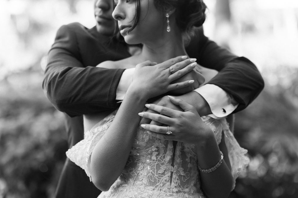 Black and white photo of a romantic embrace between newlyweds, with close-up detail of lace wedding dress and hands.