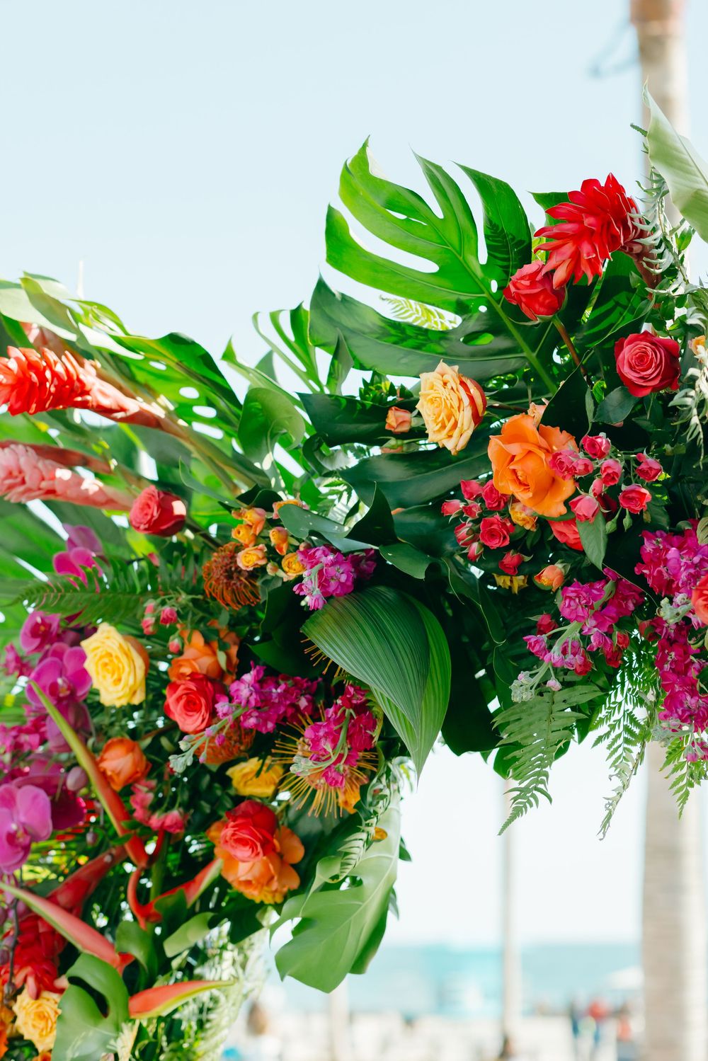 Vibrant tropical floral arrangement with red roses, orange blooms and monstera leaves against a beachy background.