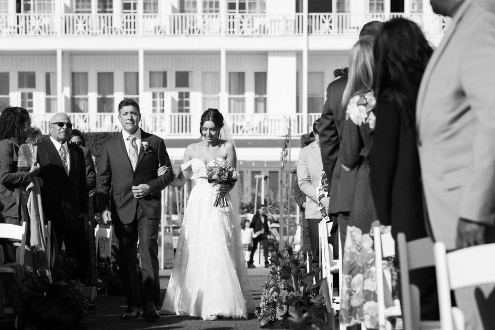 A black and white photo of a wedding processional walking down an outdoor aisle with guests seated on either side.